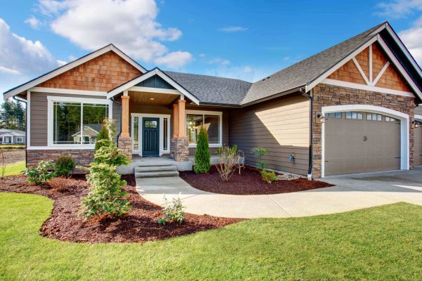 Brown craftsman style home exterior, green landscaping with shrubs, natural wood posts on porch, and dark teal door.